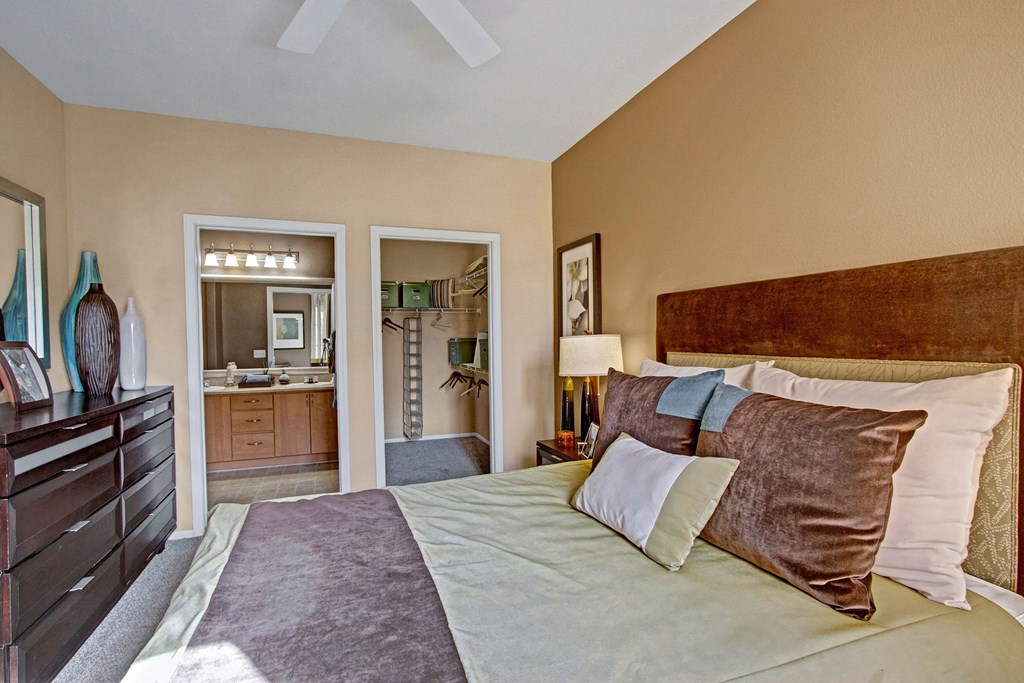 Elegant bedroom here at Fresco Apartment Homes with beige walls, a large wooden bed, and plush pillows. A dark dresser with vases is on the left, and an en suite bathroom and closet are visible.