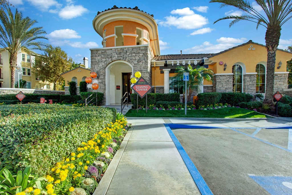 Sunny exterior of our modern leasing center here at Fresco Apartment Homes with arched entrance, surrounded by lush greenery, colorful flowers, and palm trees; welcoming atmosphere.