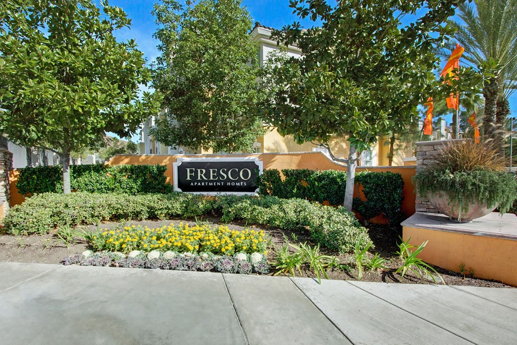Ornamental garden in front of "Fresco Apartment Homes" sign. Lush greenery and yellow flowers border the sign, creating a welcoming atmosphere.