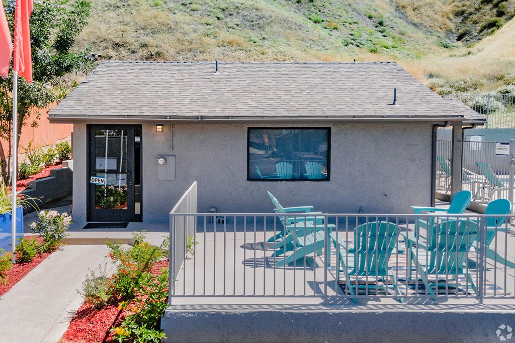 Small building here at Hillside Village Apartment Homes with a "Open" sign on the door, set against a grassy hill. Patio has turquoise chairs, creating a welcoming and relaxed atmosphere.