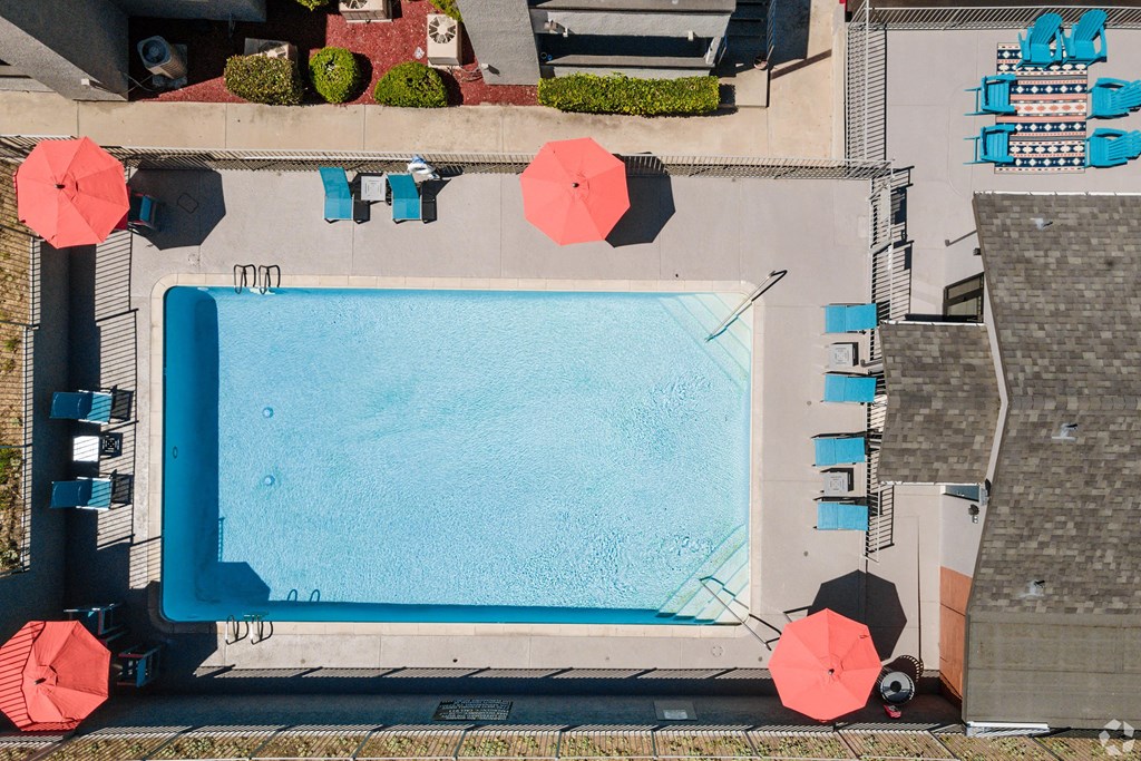 Aerial view of a rectangular swimming pool here at Hillside Village Apartment Homes surrounded by red umbrellas and blue lounge chairs. There's a patterned outdoor rug and a relaxed, inviting atmosphere.