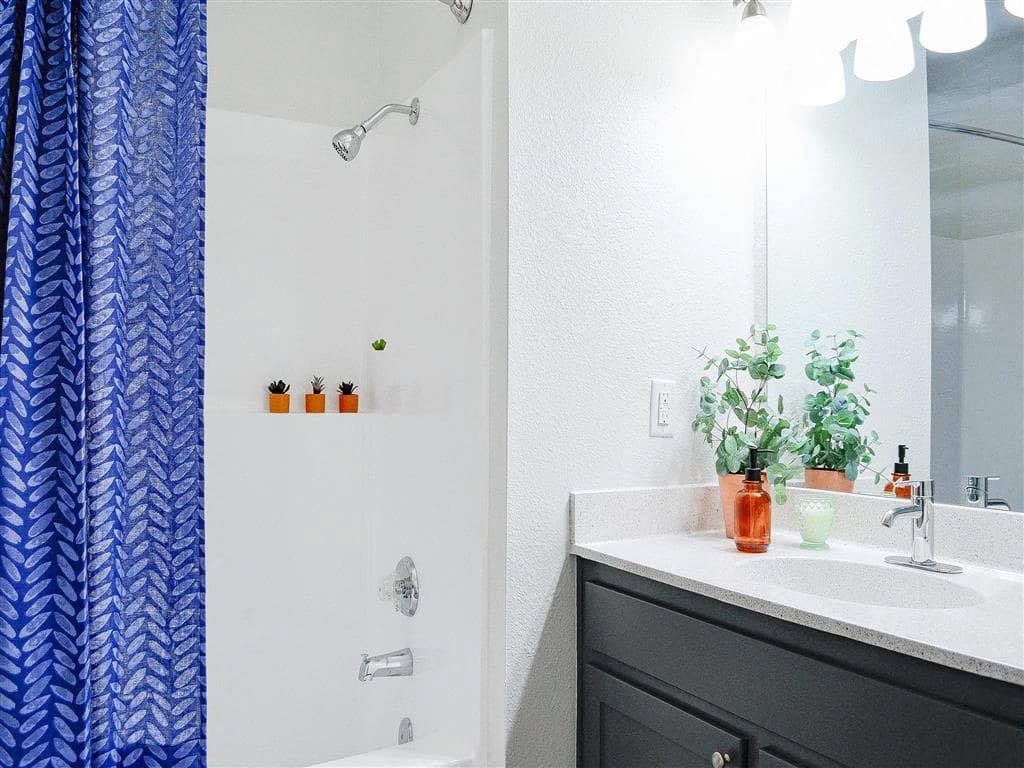Bright modern bathroom here at Hillside Village Apartment Homes with blue leafy shower curtain, white countertop, green plant in orange pot, and amber soap dispenser beside the sink.