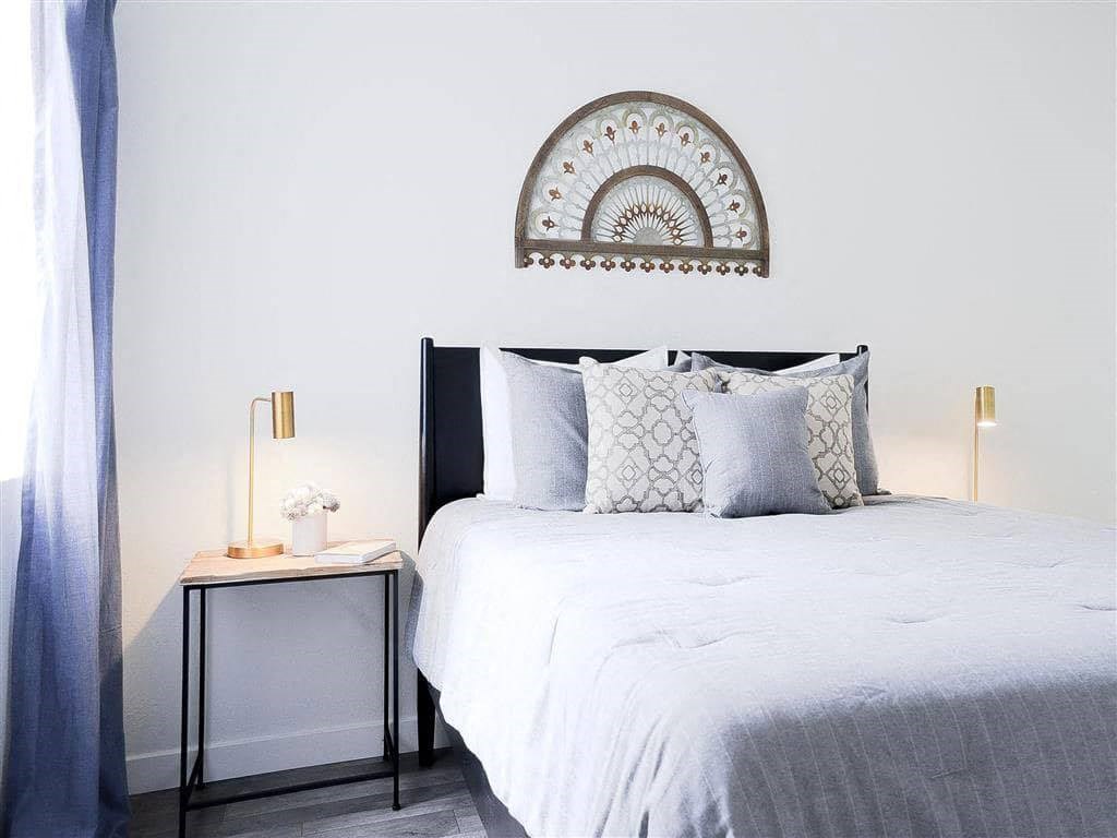 Minimalist bedroom here at Hillside Village Apartment Homes with a neatly-made bed featuring gray and patterned pillows. A semi-circular decorative wall piece hangs above. Soft lighting from two brass lamps on each side adds warmth. A small side table with a vase and book is on the left, beside sheer blue curtains.