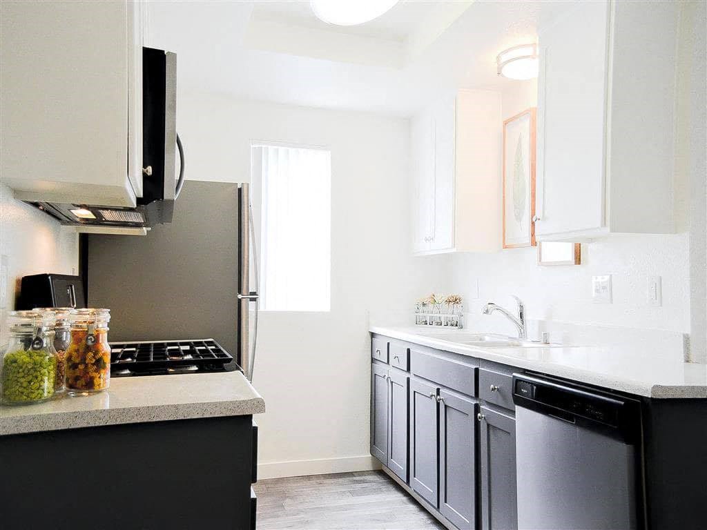 Bright modern kitchen here at Hillside Village Apartment Homes with white and gray cabinetry, stainless steel appliances, and jars of spices. Sunlight filters through a slim window, creating a clean and inviting atmosphere.