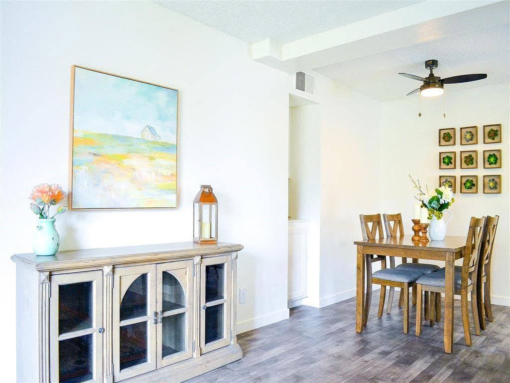 Bright dining room here at Hillside Village Apartment Homes with wooden furniture. A sideboard holds a lantern and vase with flowers. Art on walls adds warmth. Ceiling fan overhead suggests coziness.