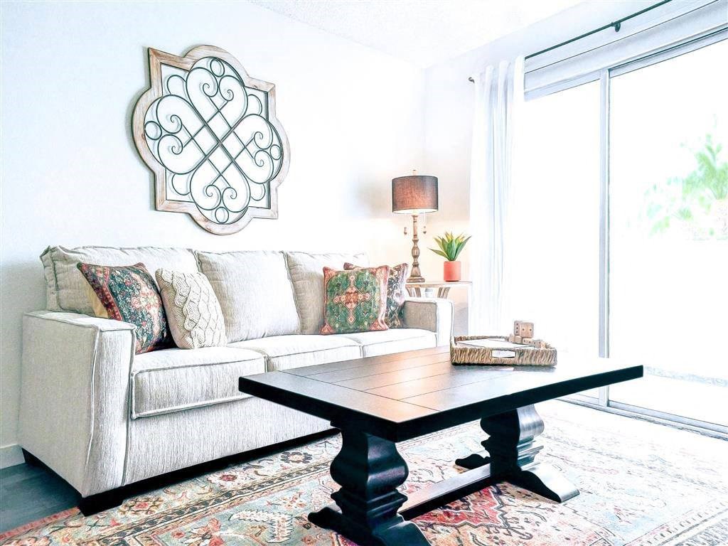 A cozy living room here at Hillside Village Apartment Homes with a beige sofa adorned with patterned cushions, a black wooden coffee table, decorative wall art, and a warm lamp beside a potted plant.