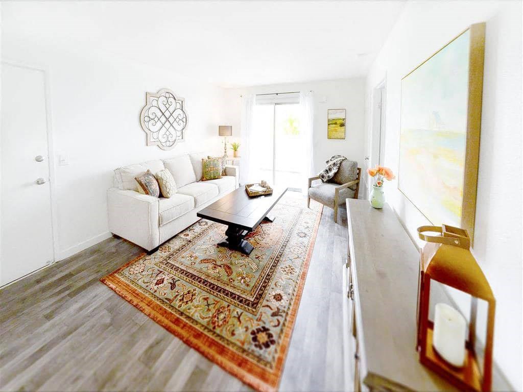 Bright living room here at Hillside Village Apartment Homes with a beige sofa, patterned rug, and wooden coffee table. Soft lighting, large painting, and sliding door create a cozy, inviting atmosphere.