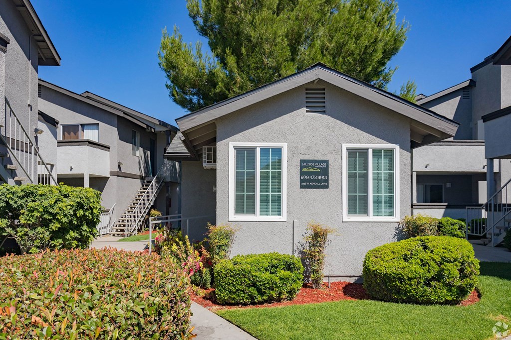 Hillside Village Apartment Homes complex with gray stucco buildings, surrounded by lush greenery and vibrant flowers. Clear blue sky creates a serene, inviting atmosphere.