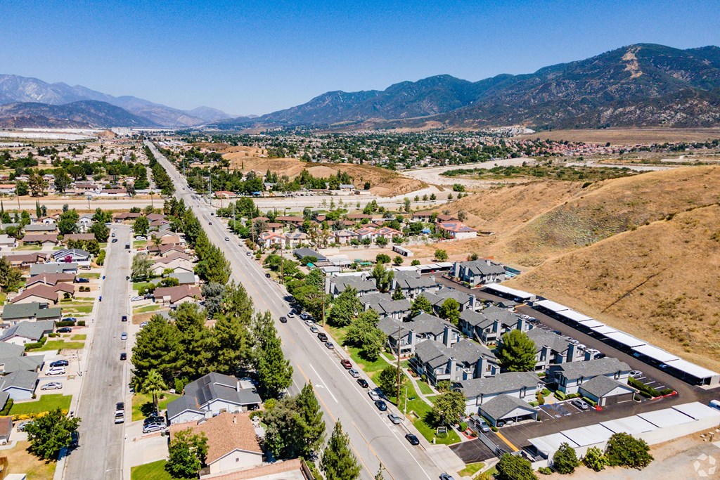 Aerial view of a suburban neighborhood near Hillside Village Apartment Homes under a clear blue sky. Lined with trees, streets divide residential homes and apartments. Hills and mountains form a scenic backdrop.