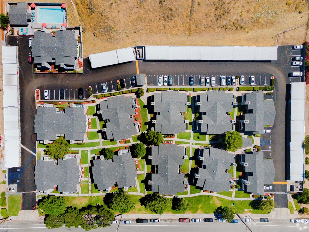 Aerial view of Hillside Village Apartment Homes featuring multiple gray-roofed buildings, green lawns, and a pool. Cars are parked in lots around the buildings.