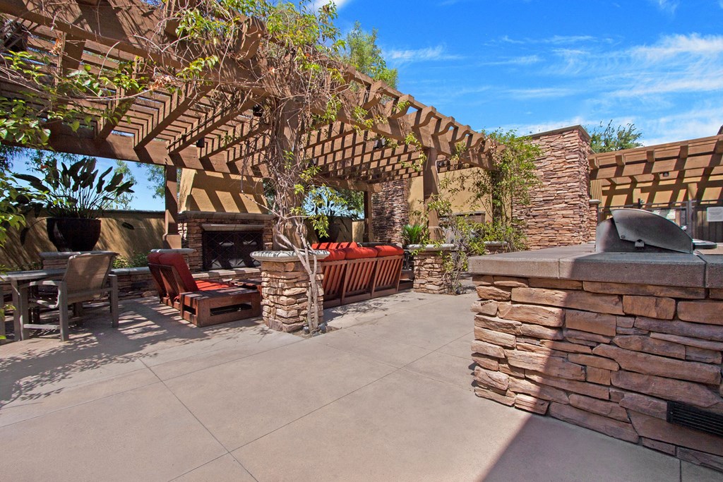Outdoor patio here at Vesada Apartment Homes with stone fireplace, red-cushioned seating, wooden pergola with climbing plants, and a built-in grill on a sunny day. Relaxing and inviting atmosphere.