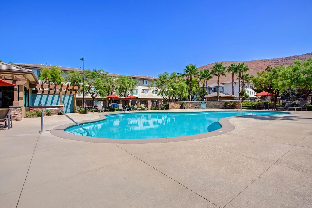 Outdoor pool here at Vesada Apartment Homes with clear blue water surrounded by palm trees and lounge chairs under red umbrellas. Residential buildings and mountains in the background. Bright, sunny day.