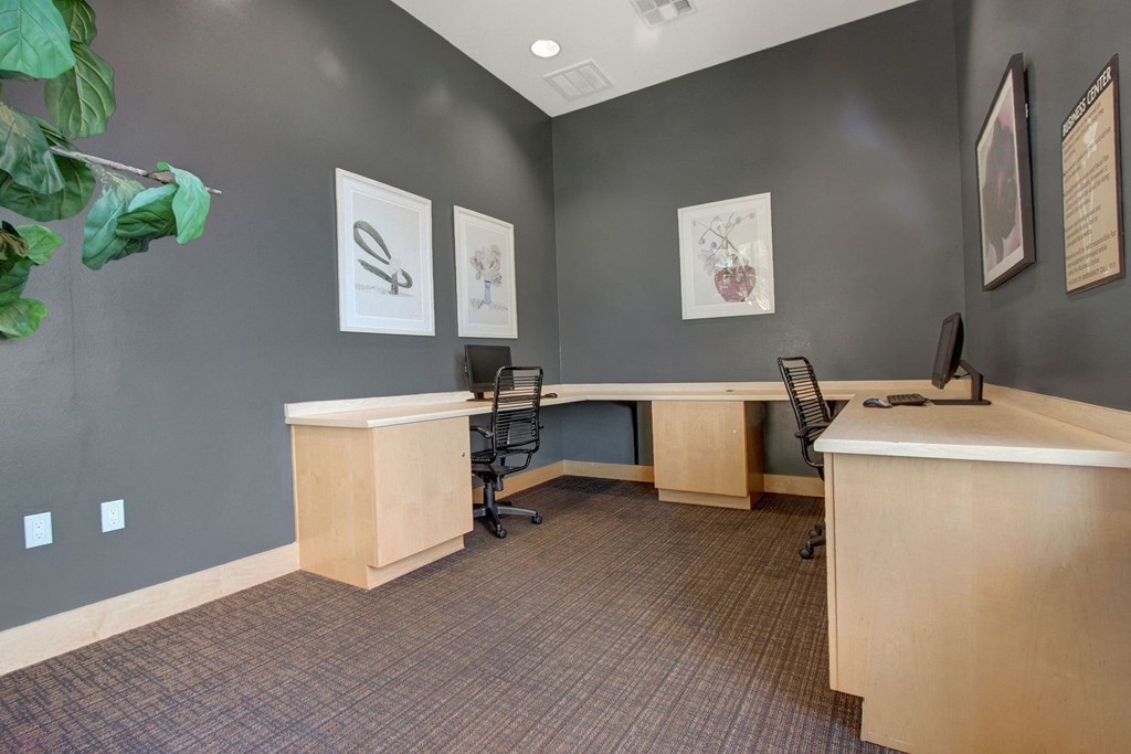 Small office space here at Vesada Apartment Homes with gray walls, two wooden L-shaped desks, black chairs, and computers. Framed artwork decorates the walls, and a plant adds a touch of greenery.