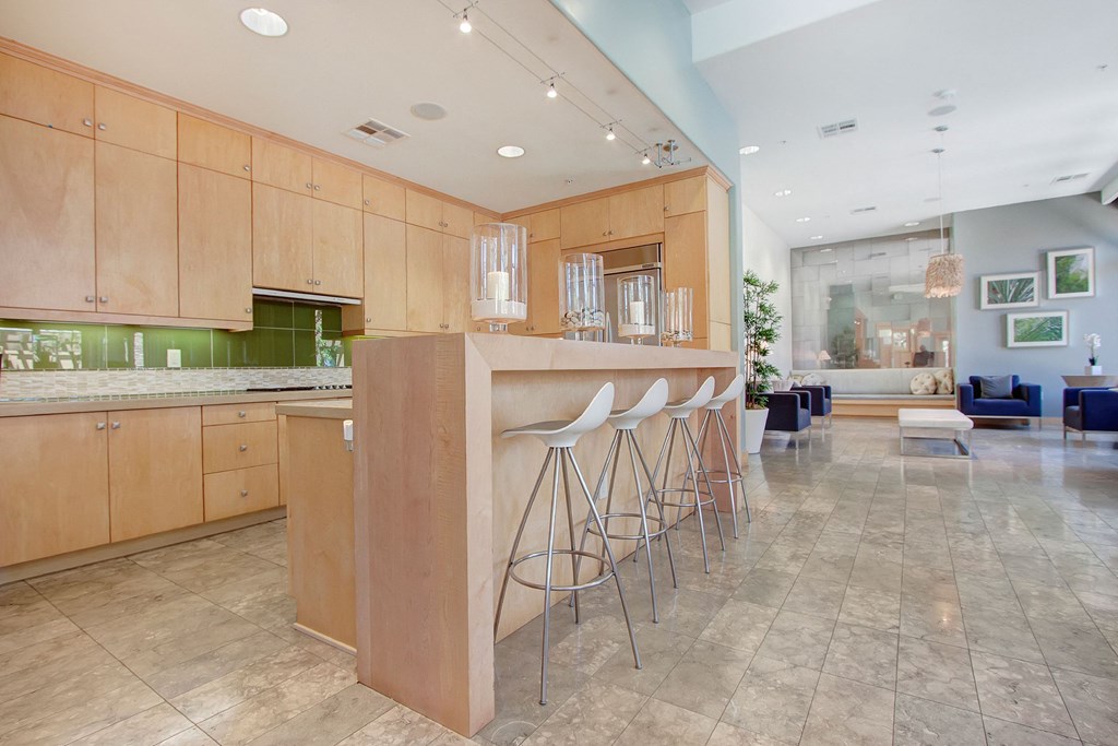 Spacious, modern resident kitchen here at Vesada Apartment Homes features light wood cabinets, a sleek island with four white bar stools. Adjacent lounge area has blue chairs and decor.