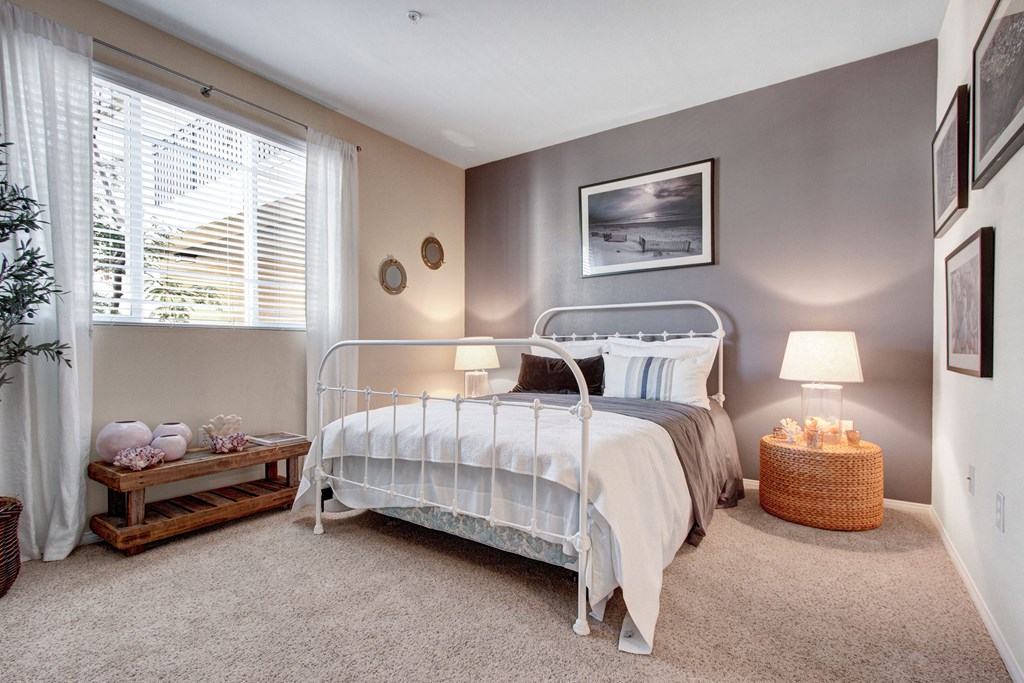 Cozy bedroom here at Vesada Apartment Homes with a white iron bed, neutral bedding, gray accent wall, and soft lighting from lamps. A window with sheer curtains adds natural light.