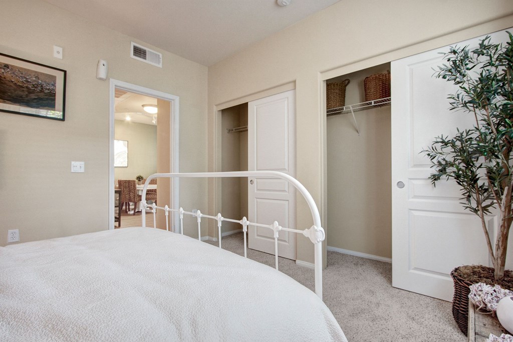 A cozy bedroom here at Vesada Apartment Homes with a white metal bed, white bedding, and a partially open closet with woven baskets. A green plant adds a touch of nature. Warm tones.