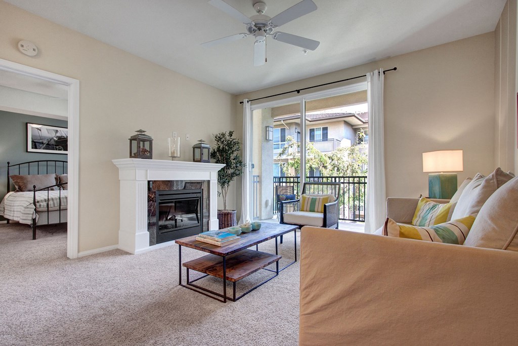 Bright living room here at Vesada Apartment Homes with beige walls, a cozy sofa, and colorful pillows. It features a white fireplace, wooden coffee table, and large window opening to a balcony. A bedroom is visible through an open door, creating a welcoming ambiance.