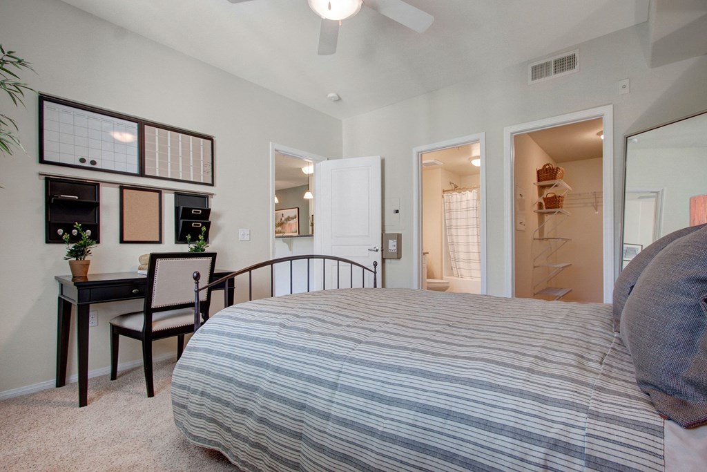 Neutral-toned bedroom here at Vesada Apartment Homes with striped bedding, black desk, and wall organizers. Open doors reveal a bathroom and a closet. Calm and organized atmosphere.