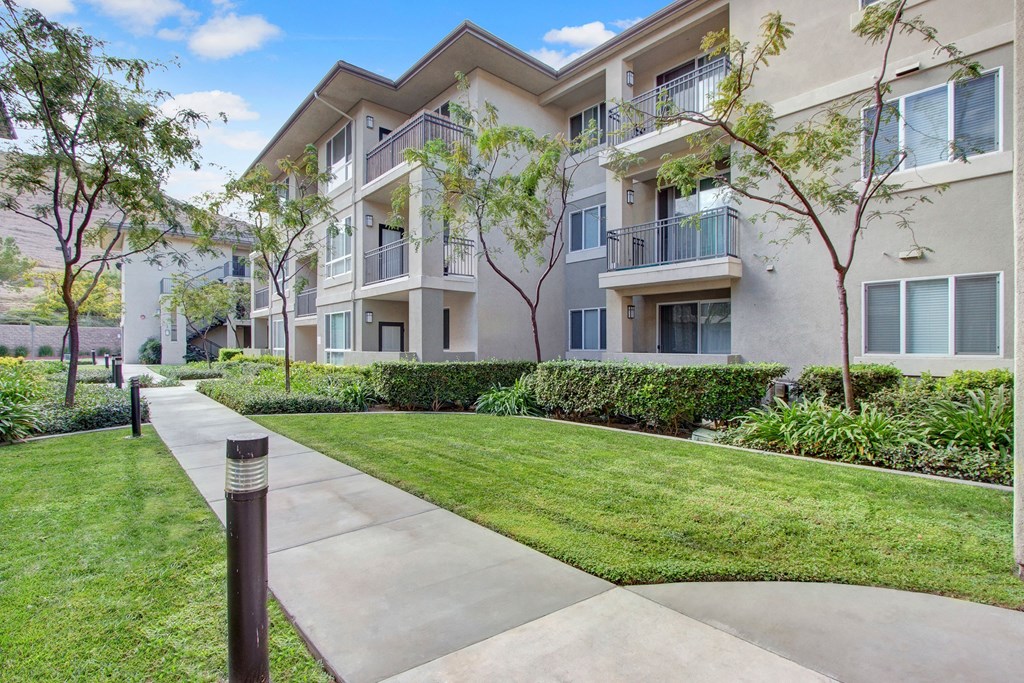 Modern three-story Vesada Apartment Homes building with balconies, surrounded by lush greenery and trees. A pathway leads through the well-maintained lawn, under a bright blue sky.