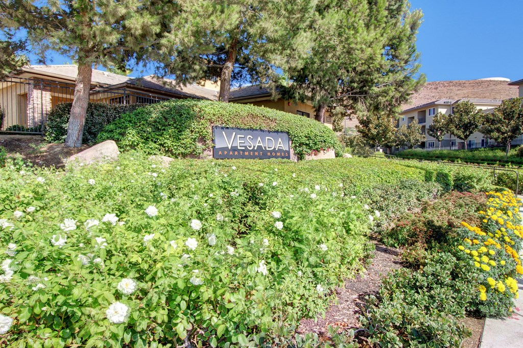 Lush garden with white and yellow flowers surrounds the "Vesada Apartment Homes" sign, framed by tall green trees and buildings in the background.