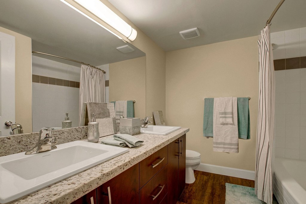 Modern bathroom here at 3600 Brentwood Apartment Homes with dual sinks on a granite countertop, wood cabinets, a shower with a striped curtain, and soft beige walls. Calm and sleek ambiance.