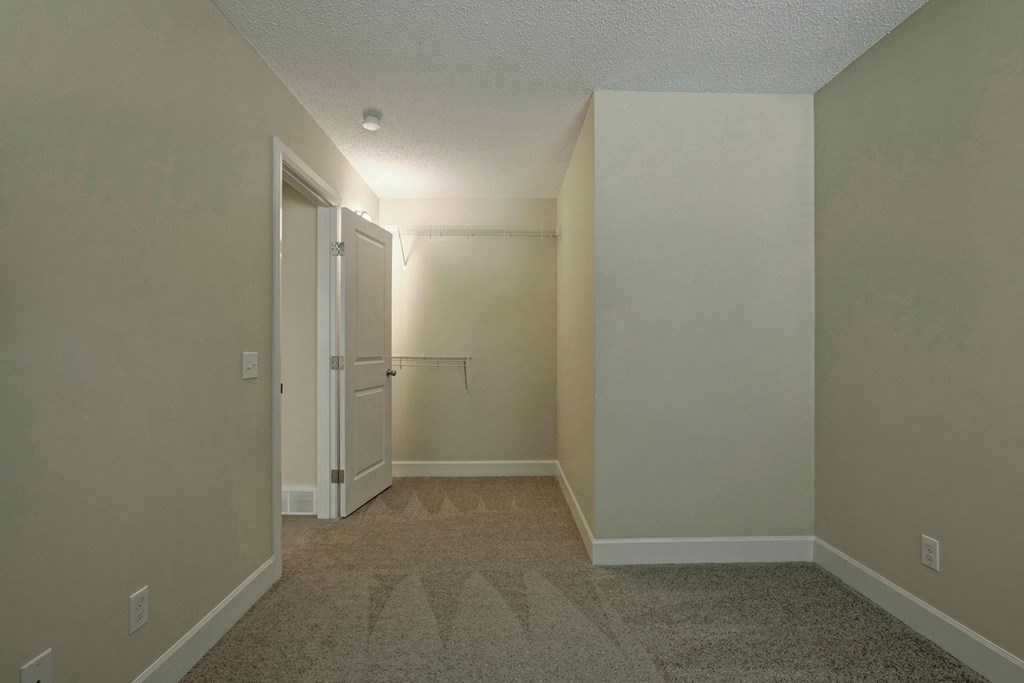 Empty bedroom here at 3600 Brentwood Apartment Homes with beige walls, an open white door, and a small closet with a wire shelf. The carpet is gray, creating a calm, neutral tone.