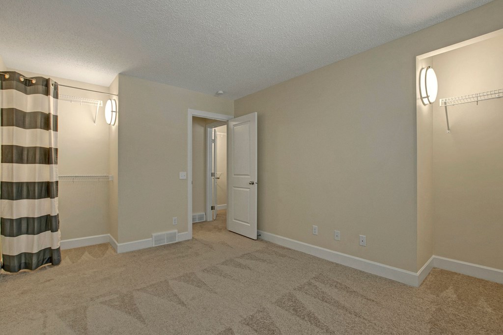 Empty beige bedroom here at 3600 Brentwood Apartment Homes with carpeted floor, open door, and striped curtains. Wall sconces illuminate shelves on each side, creating a neutral, calm ambiance.