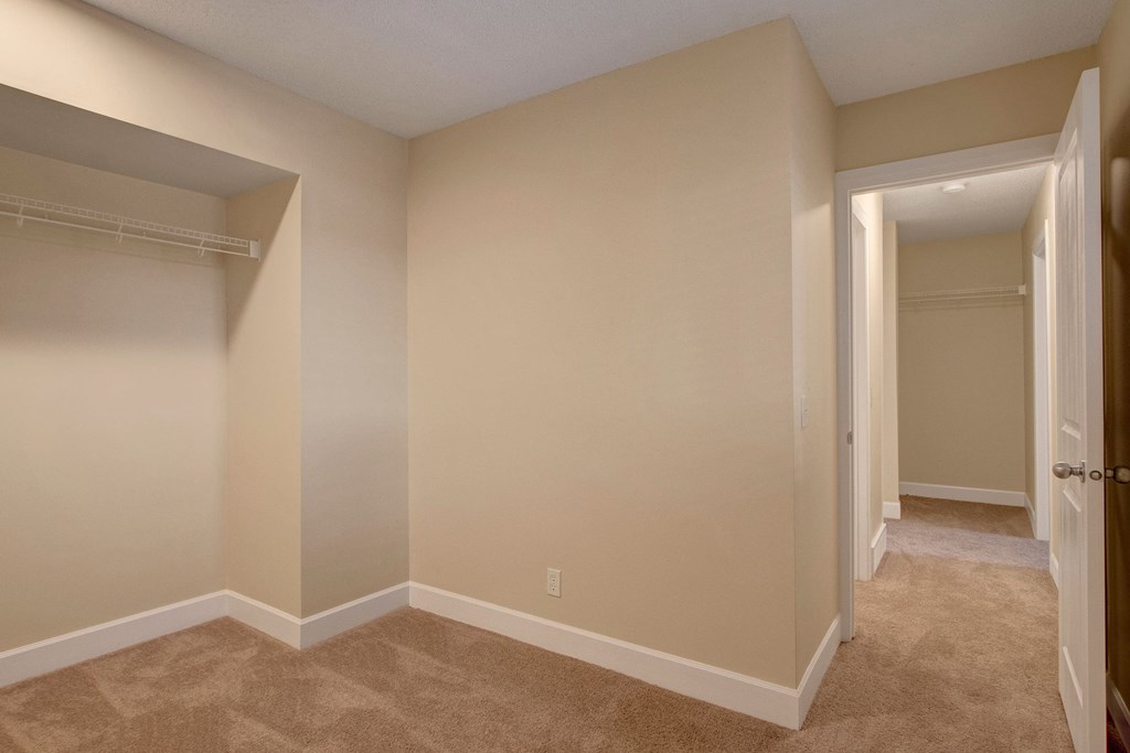 Empty beige bedroom here at 3600 Brentwood Apartment Homes with beige carpet and open closet. A small hallway leads to another similar room. Calm, neutral atmosphere.