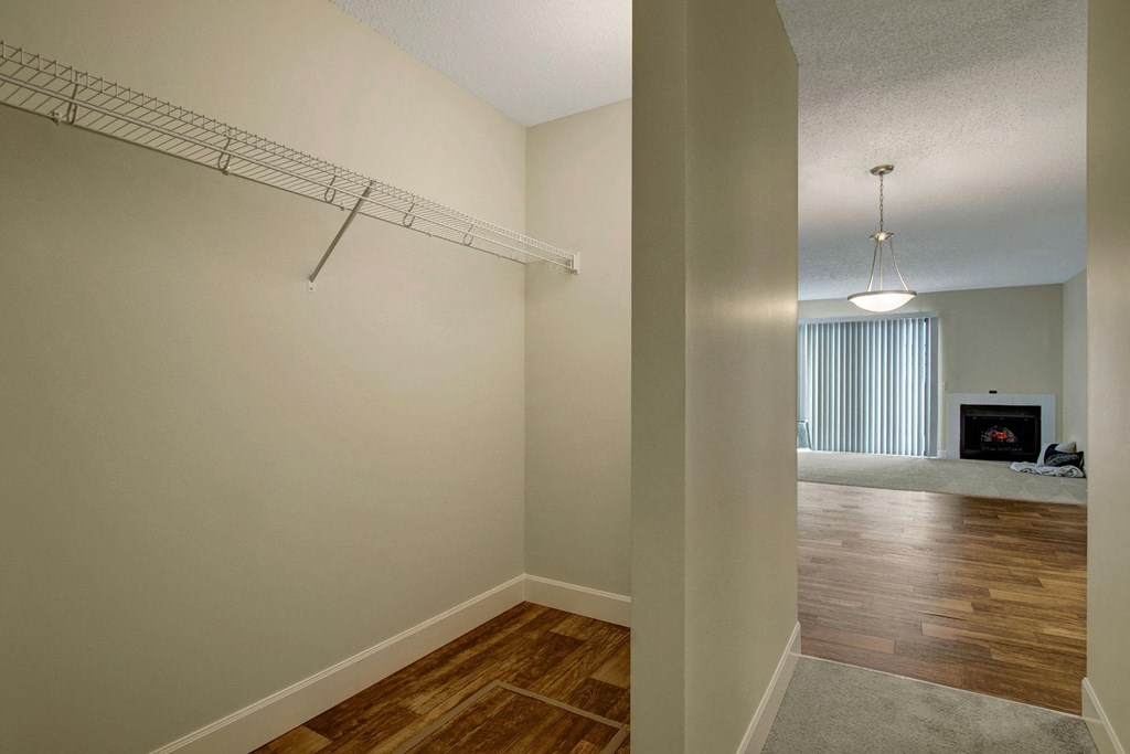 A spacious, empty walk-in closet here at 3600 Brentwood Apartment Homes with a single wire shelf. Adjacent room features wooden flooring, a lit pendant lamp, vertical blinds, and a cozy fireplace.