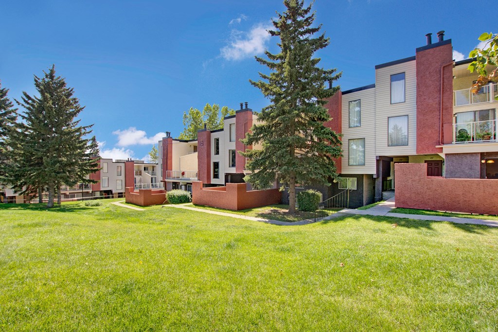 Modern 3600 Brentwood Apartment Homes complex with red and beige buildings surrounded by lush green grass and tall pine trees under a clear blue sky. Tranquil atmosphere.