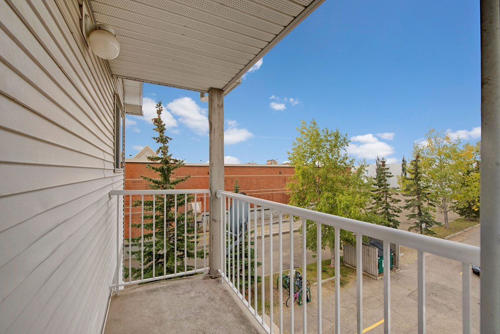 View from a small balcony here at Aspen Terrace Apartment Homes with white railing, overlooking a parking lot with bicycles, trees, and a brick building. Clear blue sky with scattered clouds.