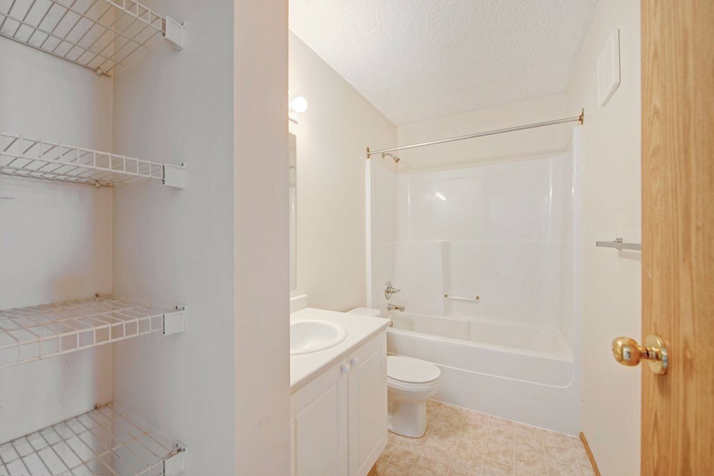 Compact bathroom here at Aspen Terrace Apartment Homes with white fixtures. Features a sink, toilet, and bathtub. Wire shelves are on the left, and a wooden door on the right. Clean and minimal.