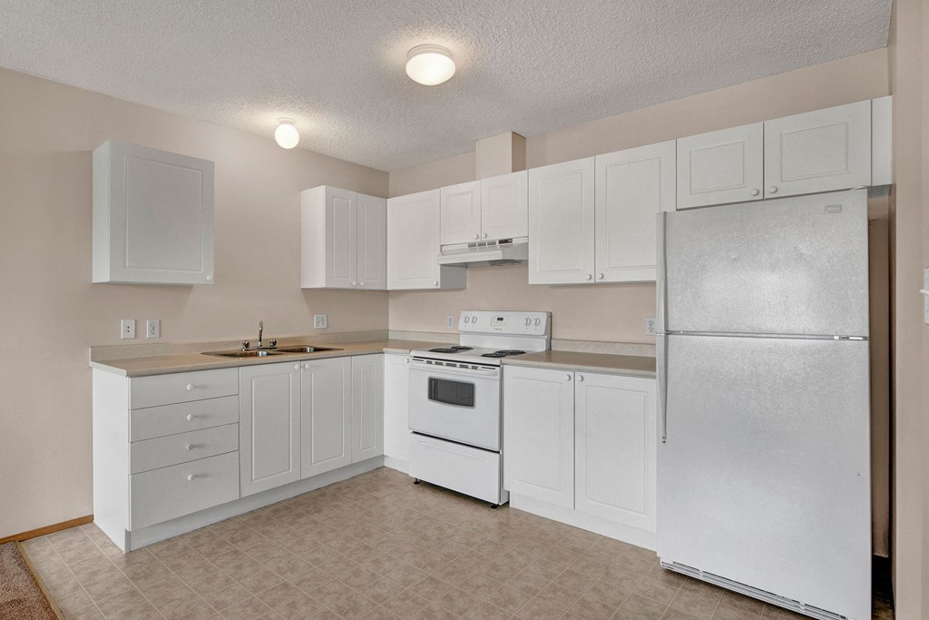 Minimalist kitchen here at Aspen Terrace Apartment Homes with white cabinets, stove, and refrigerator. Beige walls and tiled floor create an airy, neutral atmosphere. Bright lighting.