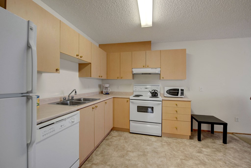A clean, compact kitchen here at Aspen Terrace Apartment Homes with light wood cabinets, white appliances, and beige countertop. Includes a fridge, stove, microwave, and dishwasher. Casual tone.