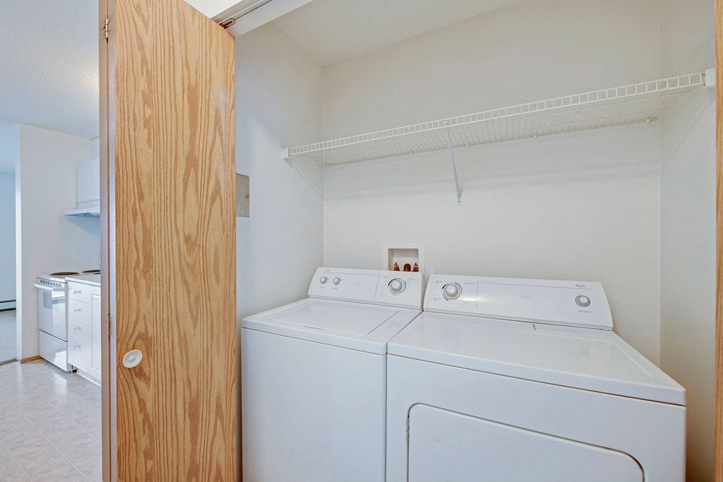 Laundry area here at Aspen Terrace Apartment Homes with white washer and dryer, enclosed by a wooden sliding door. A white wire shelf is above. Kitchen appliances are visible in the background.