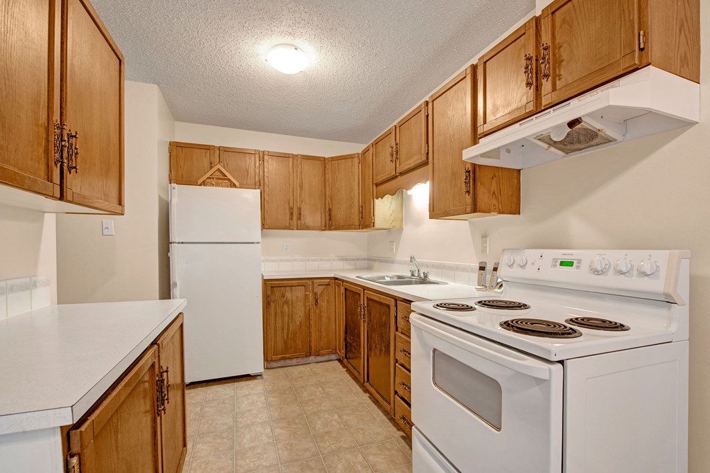 Cozy kitchen here at Broadmoor Apartment Homes with wooden cabinets, white countertops, and a tiled floor. Features a white fridge, stove, and sink beneath warm lighting.