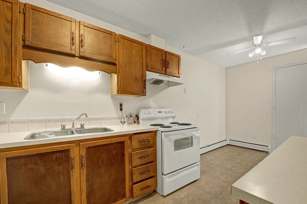 A cozy kitchen here at Broadmoor Apartment Homes featuring wooden cabinets, a double sink, a white stove, and a ceiling fan. The space radiates warmth with its earthy tones.