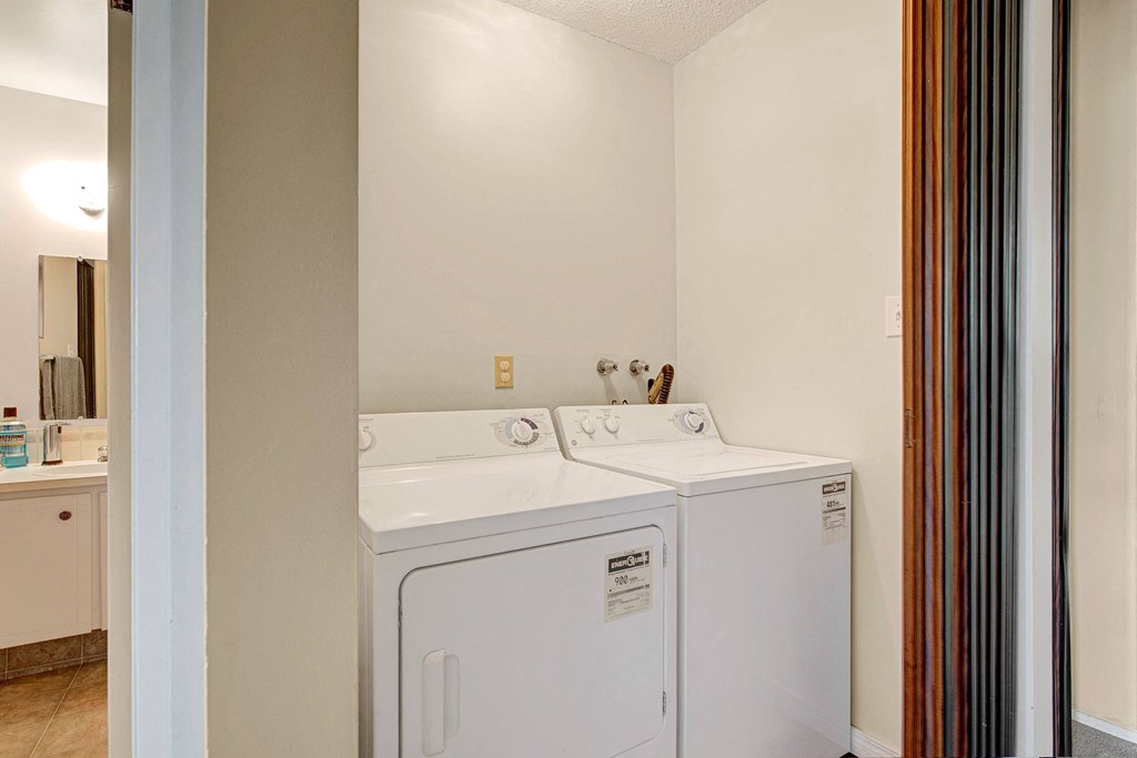 Laundry room here at Broadmoor Apartment Homes with a white washing machine and dryer side by side. To the left, a bathroom with a sink and mirror is partially visible. Soft lighting fills the space.