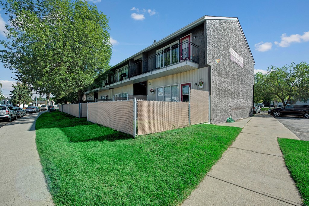 Two-story gray Broadmoor Apartment Homes building with balconies and chain-link fence, a lush green lawn, and a large tree. "Now Leasing" sign visible. Bright, clear day.