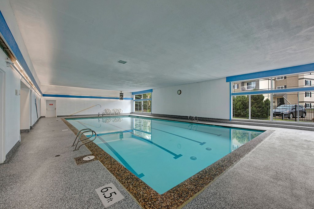 An indoor swimming pool here at Cambridge West Apartment Homes with clear blue water, surrounded by a gray tile floor. Large windows on the right let in natural light, reflecting off the pool.