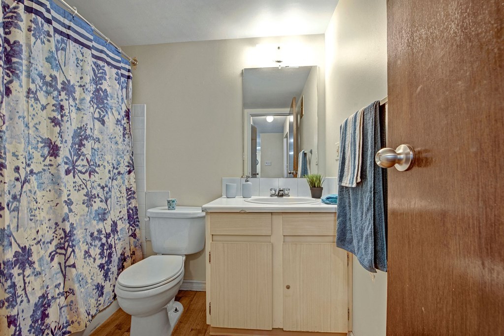 Small bathroom here at Cambridge West Apartment Homes with wooden door, floral shower curtain, toilet, and light wood vanity. A mirror above the sink reflects a cozy, simple decor.