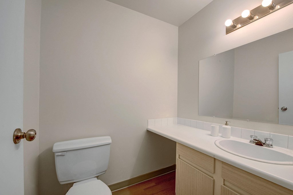 Small, clean bathroom here at Cambridge West Apartment Homes with beige walls, wooden vanity, white countertop, sink, mirror, and toilet. Soft lighting and minimal decor create a simple, calm atmosphere.