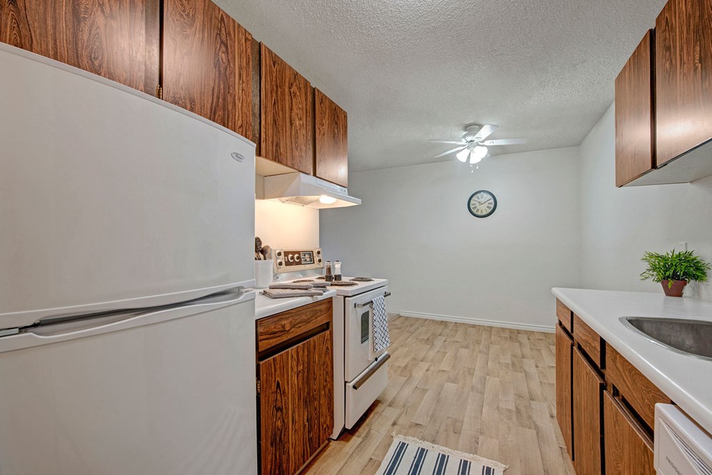 Small kitchen here at Cambridge West Apartment Homes with white appliances, wood cabinets, and light wood flooring. A ceiling fan and wall clock are visible. The space feels simple and cozy.