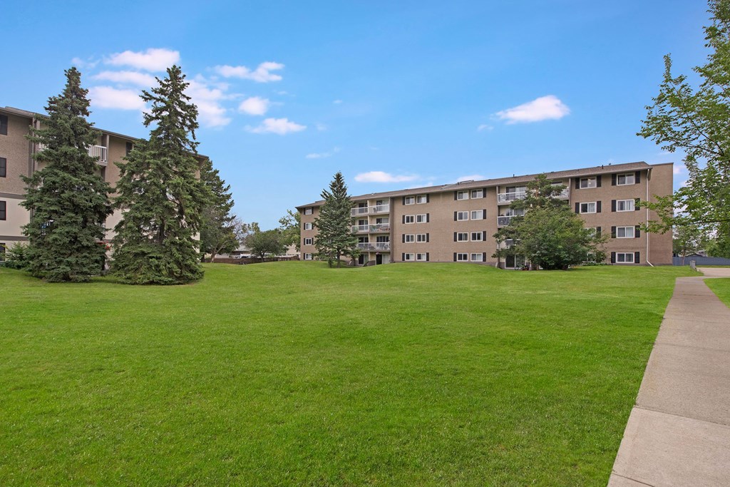 Expansive green lawn with scattered evergreen trees leads to a four-story Cambridge West Apartment Homes building under a clear blue sky, conveying tranquility and openness.