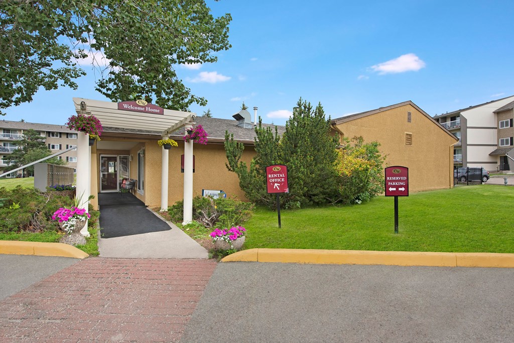 Entrance to Cambridge West Apartment Homes leasing office with a sign reading "Welcome Home" under a pergola adorned with hanging flowers. Nearby, signs indicate rental office and parking.