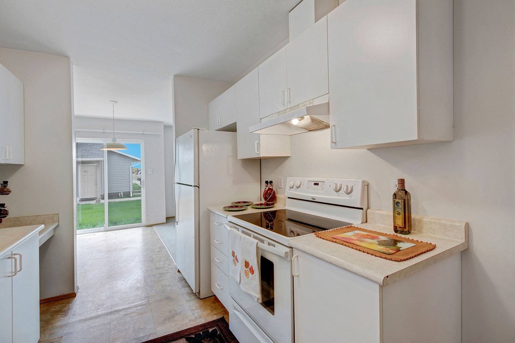 Compact kitchen here at Carrington Place Apartment Homes with white cabinets, stove, and fridge. Sunlit space via glass door to yard. Cozy, clean ambiance with minimal decor.