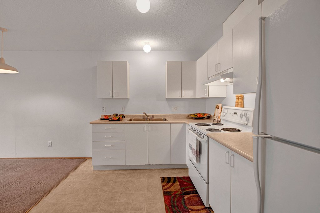 A living room with easy access to the kitchen Simple kitchen here at Carrington Place Apartment Homes with white cabinets, beige countertops, and white appliances, including a stove and fridge. A colorful rug and bowls add warmth.