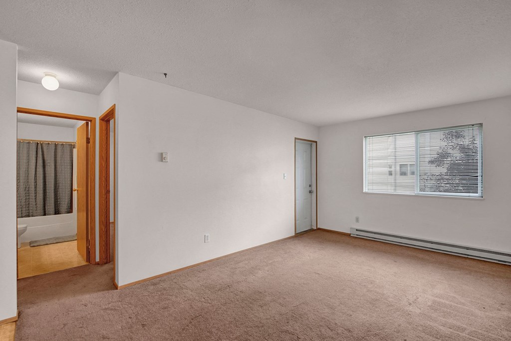 Large, bright, and comfortable living room Empty bedroom here at Carrington Place Apartment Homes with beige carpet and white walls. A window with blinds lets in soft daylight. Open door reveals a bathroom with a shower curtain. Minimalist tone.