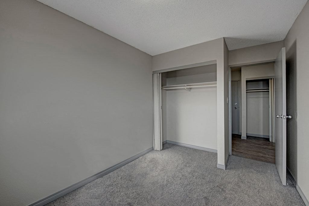 Empty bedroom here at Cedar Gardens Apartment Homes with gray walls and carpet, featuring an open closet with hanging rods. Doorway leads to hardwood floor hallway, creating a minimalist, neutral tone.