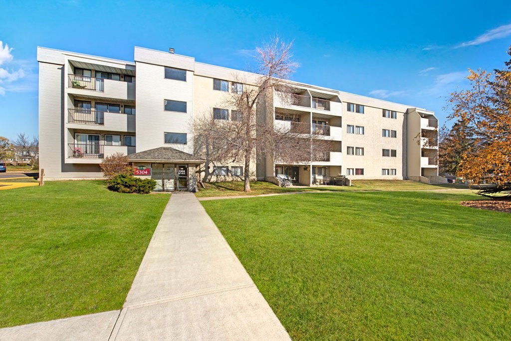 Four-story Cedar Gardens Apartment Homes building with balconies and a lawn, under a clear blue sky. A pathway leads to the entrance, creating a welcoming atmosphere.